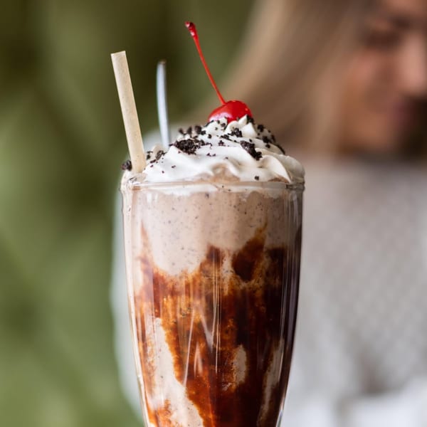 A vegan cookies and cream milkshake with whipped cream, cookie crumbles, and a cherry on top.