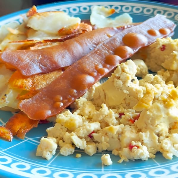 A blue plate holds vegan scrambled eggs, crispy home fries, and two strips of vegan bacon.