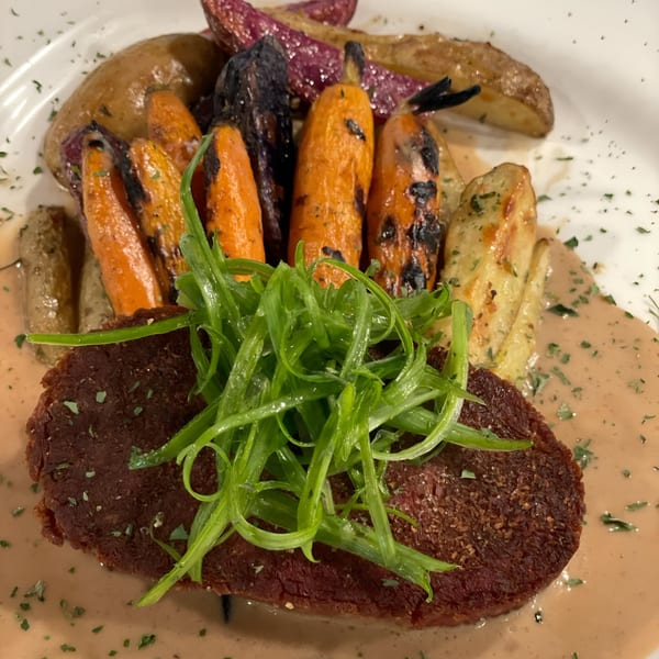Vegan steak with green onions, roasted carrots, and potatoes, drizzled with sauce.